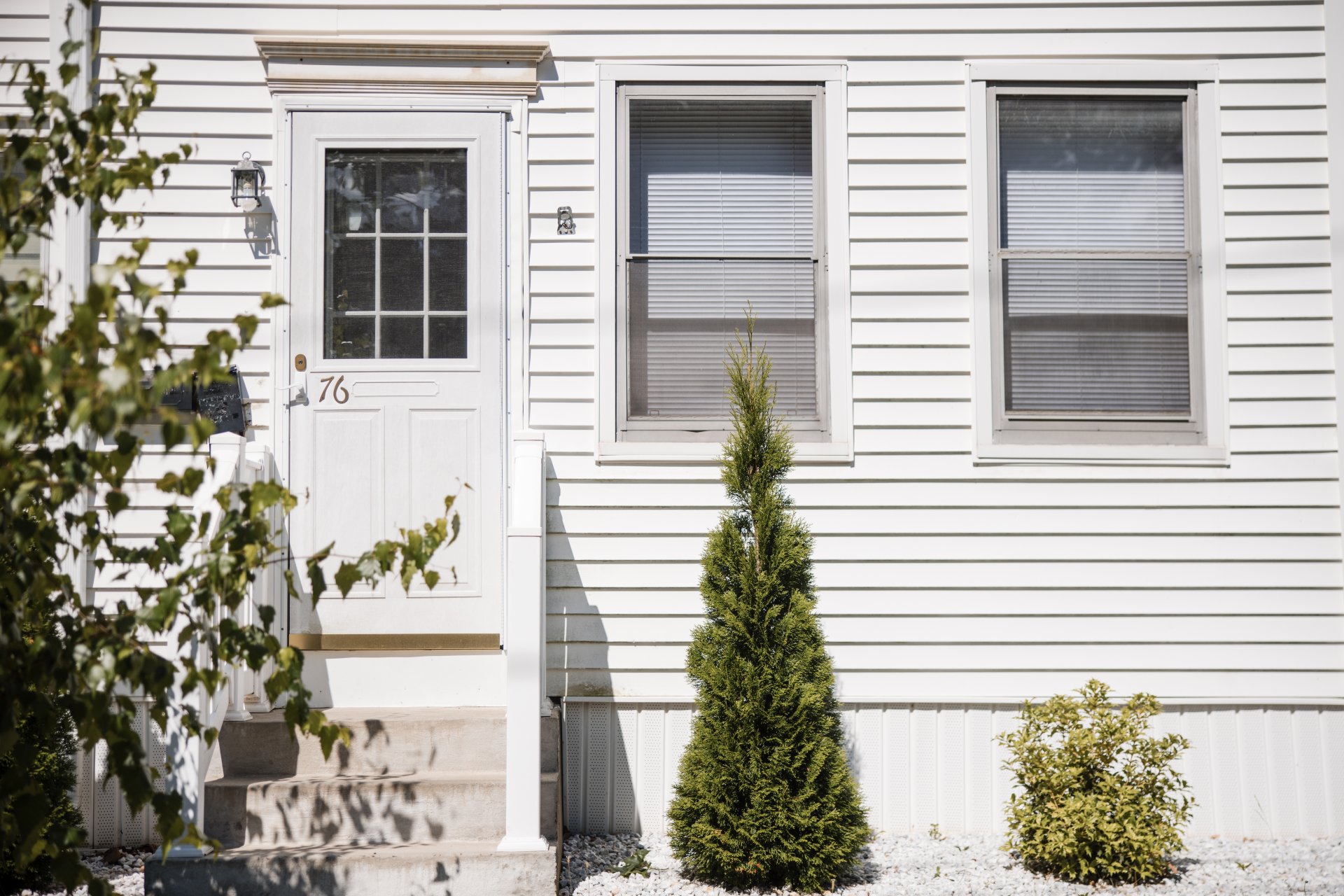  Front view of a white house with a door and two windows. A small tree and a shrub are planted near the entrance. House number 76 is displayed on the door.