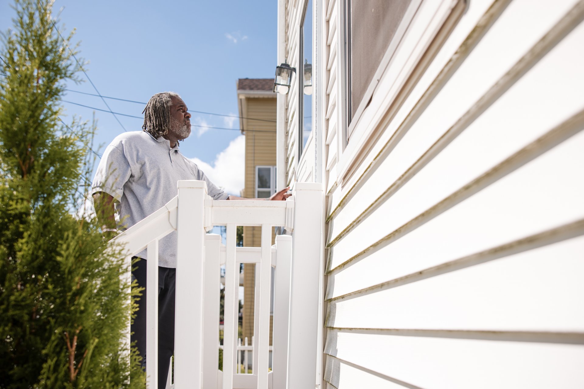  Man with gray hair standing on porch steps, looking at the house siding, with a tree on the left and another house in the background.