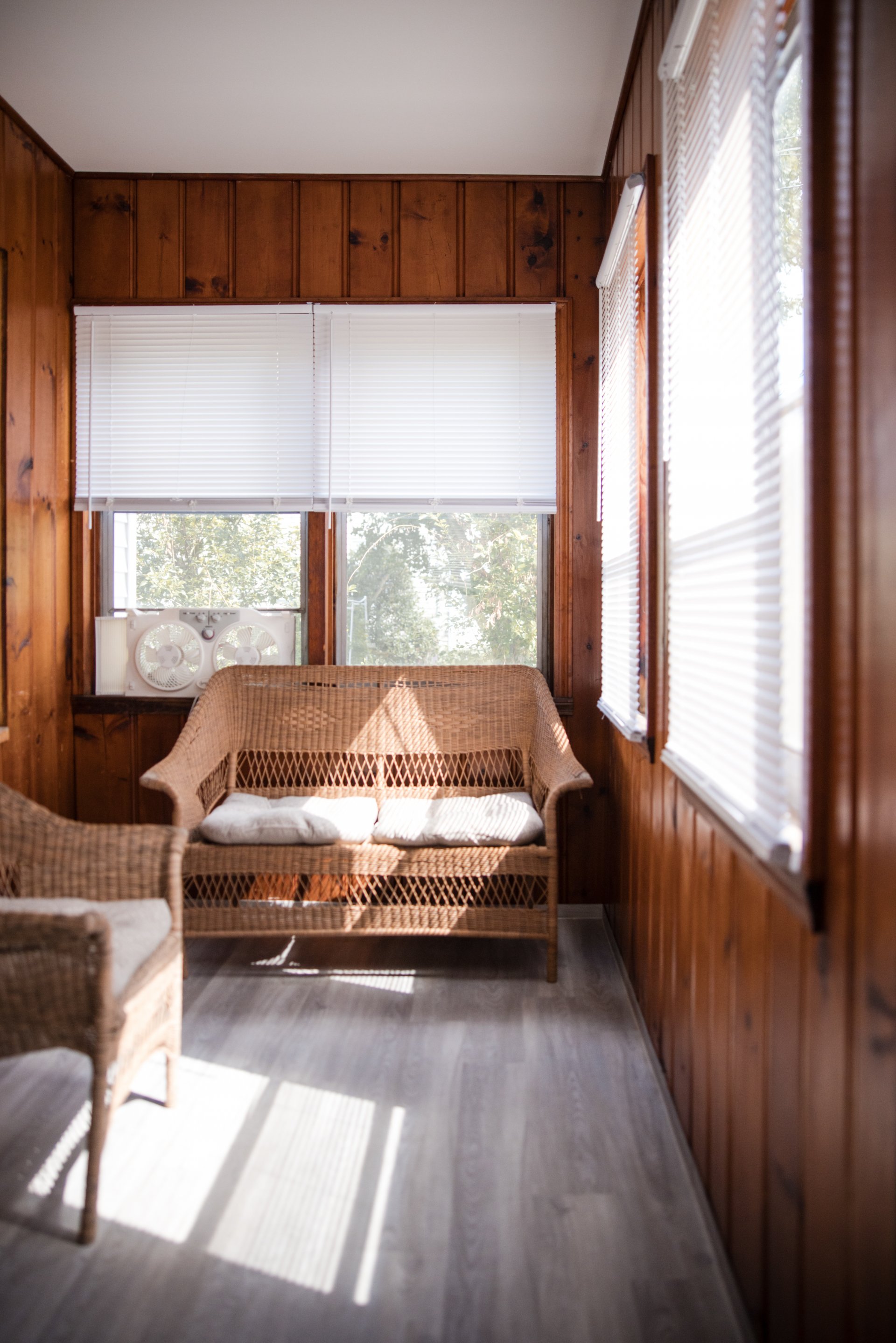  A sunlit room with wooden walls features wicker furniture and a window with blinds, letting in natural light.