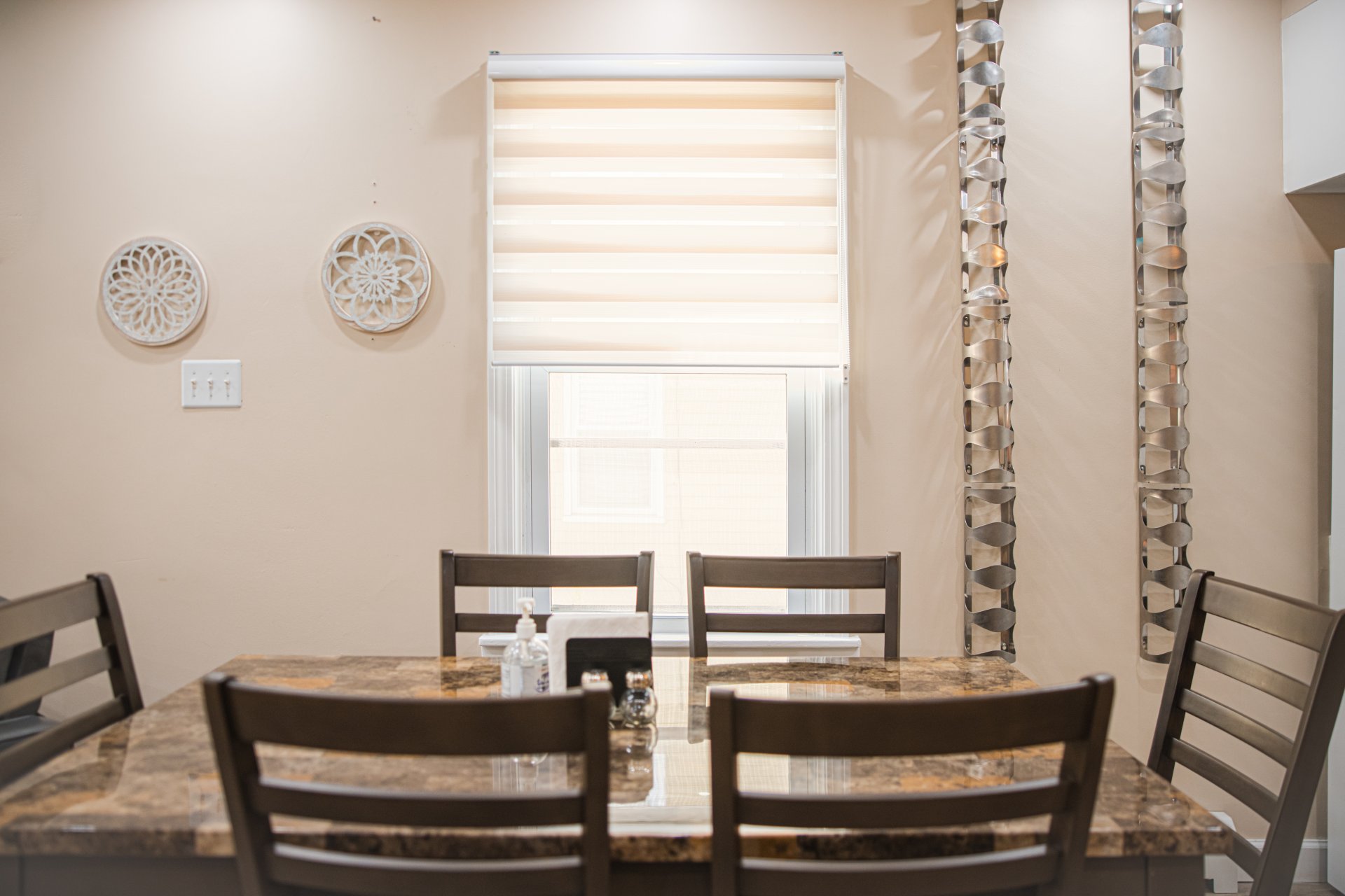  Dining area with a marble table, four chairs, and a window with a roller blind. Decorative wall hangings and metallic wall art are visible.