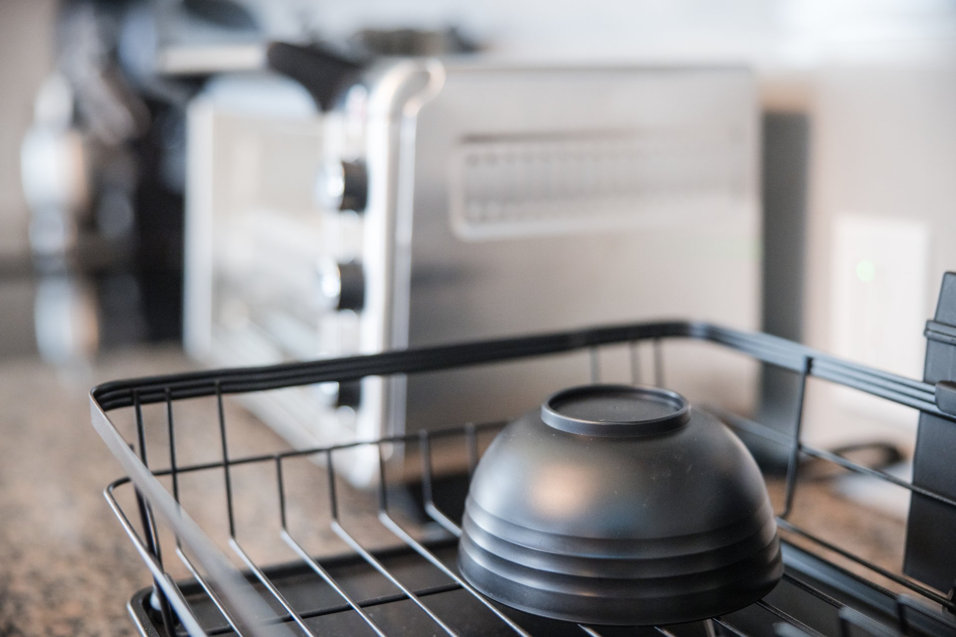  A black bowl sits upside down on a dish rack in a kitchen. In the background, a toaster oven is visible on the counter.