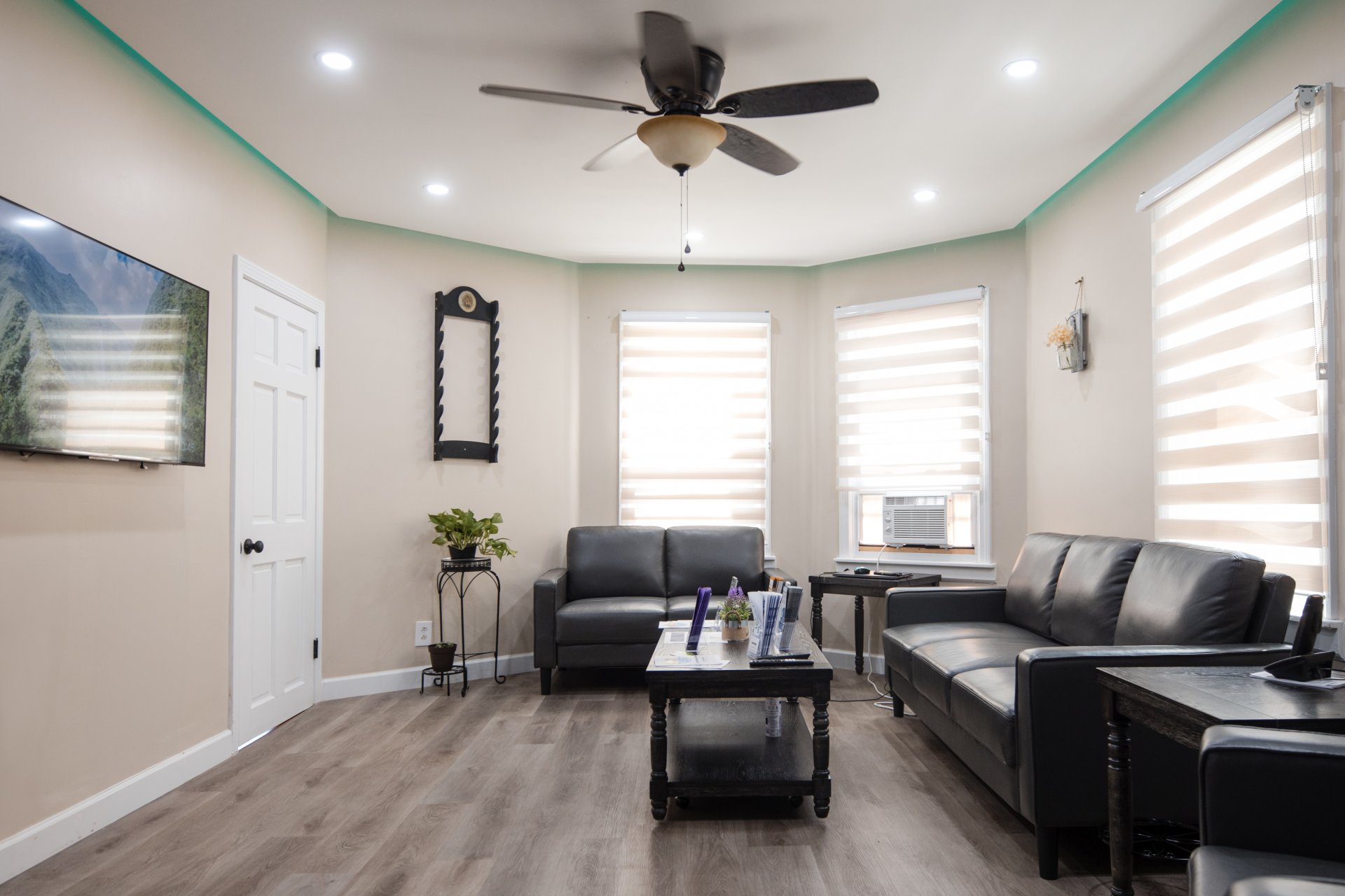 A modern living room with two black sofas, a coffee table, a ceiling fan, a wall-mounted TV, and a small plant. Windows have beige blinds, and the walls are light-colored.