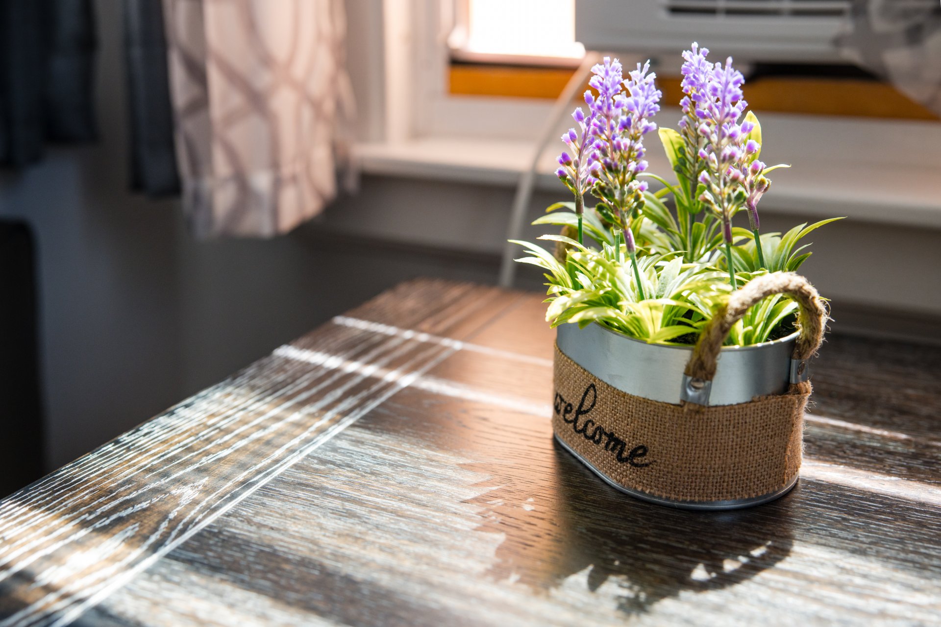  A small metal planter with lavender flowers sits on a wooden table near a sunlit window. The planter is wrapped in burlap with the word "welcome.