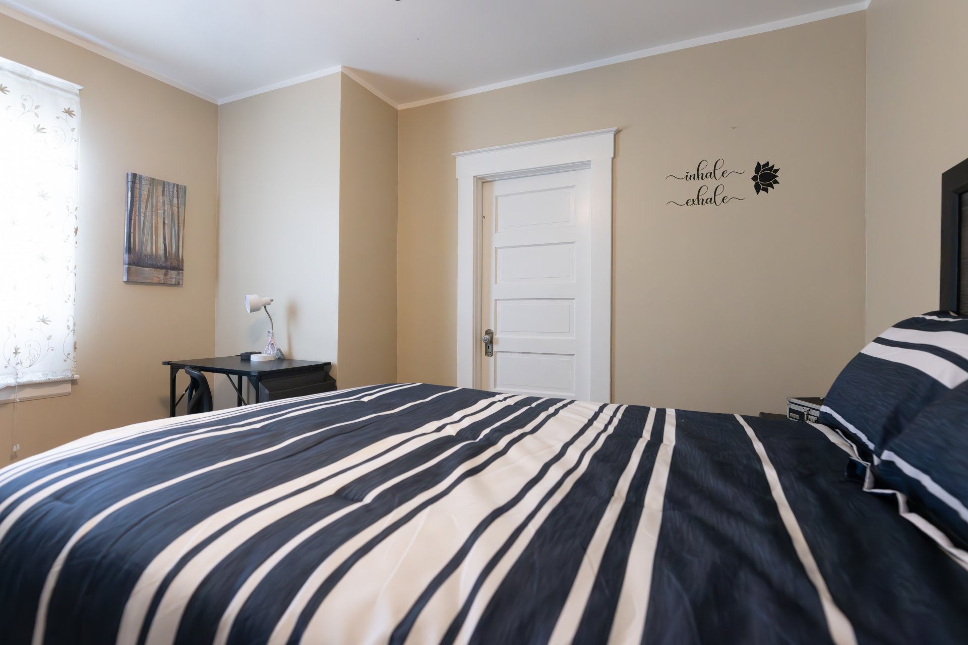  Bedroom with a bed featuring black and white striped bedding, a side table with a lamp, a window with sheer curtains, and a wall with motivational text and flower decal