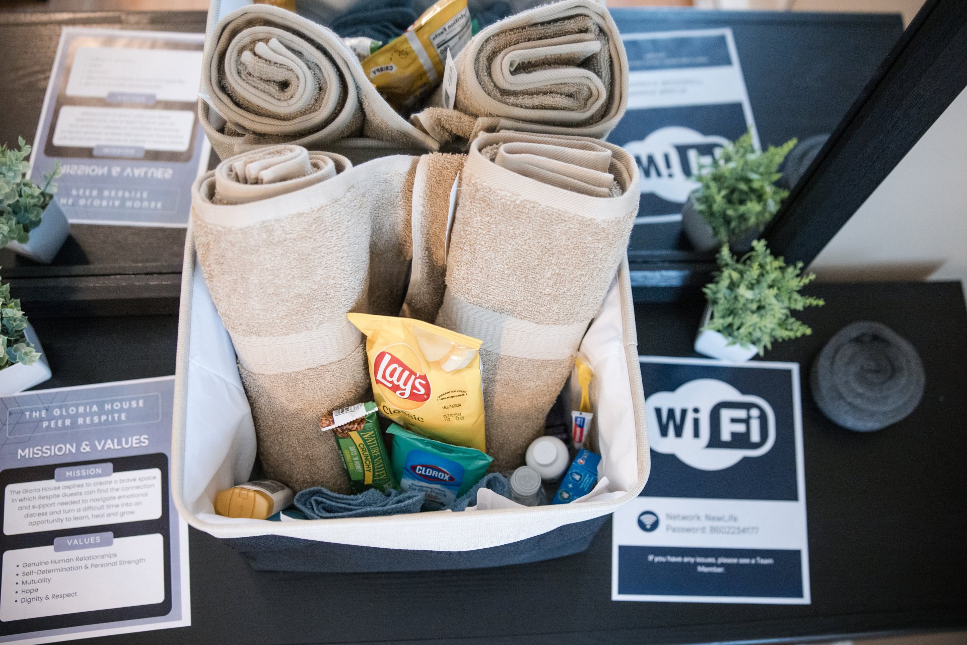  A basket holds rolled towels, a Lay's chip bag, snacks, toiletries, a WiFi sign, and a mission statement card on a counter with potted plants.