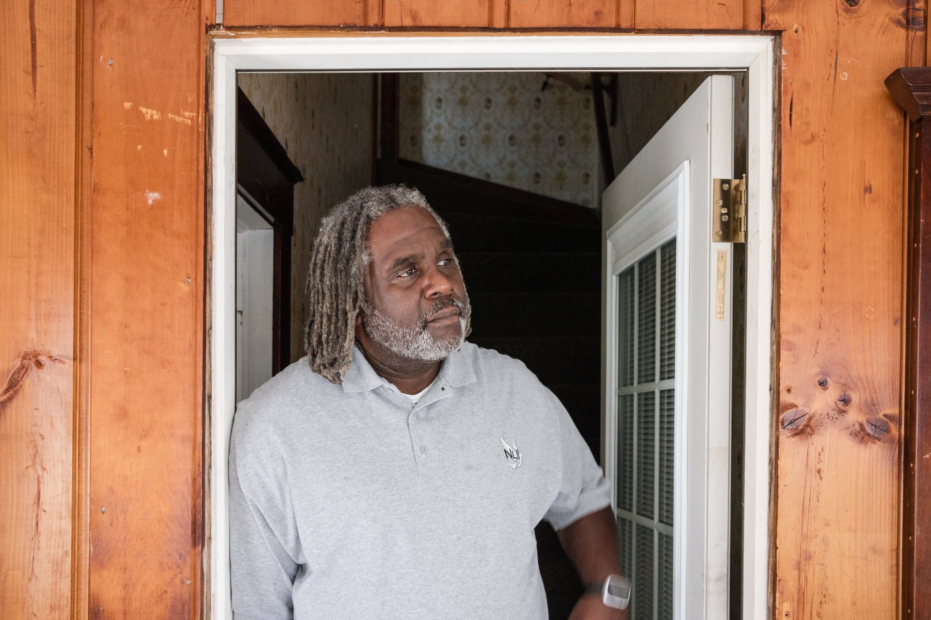  A man with grey dreadlocks and a light shirt stands in a wooden-paneled room, smiling and leaning against a doorway.