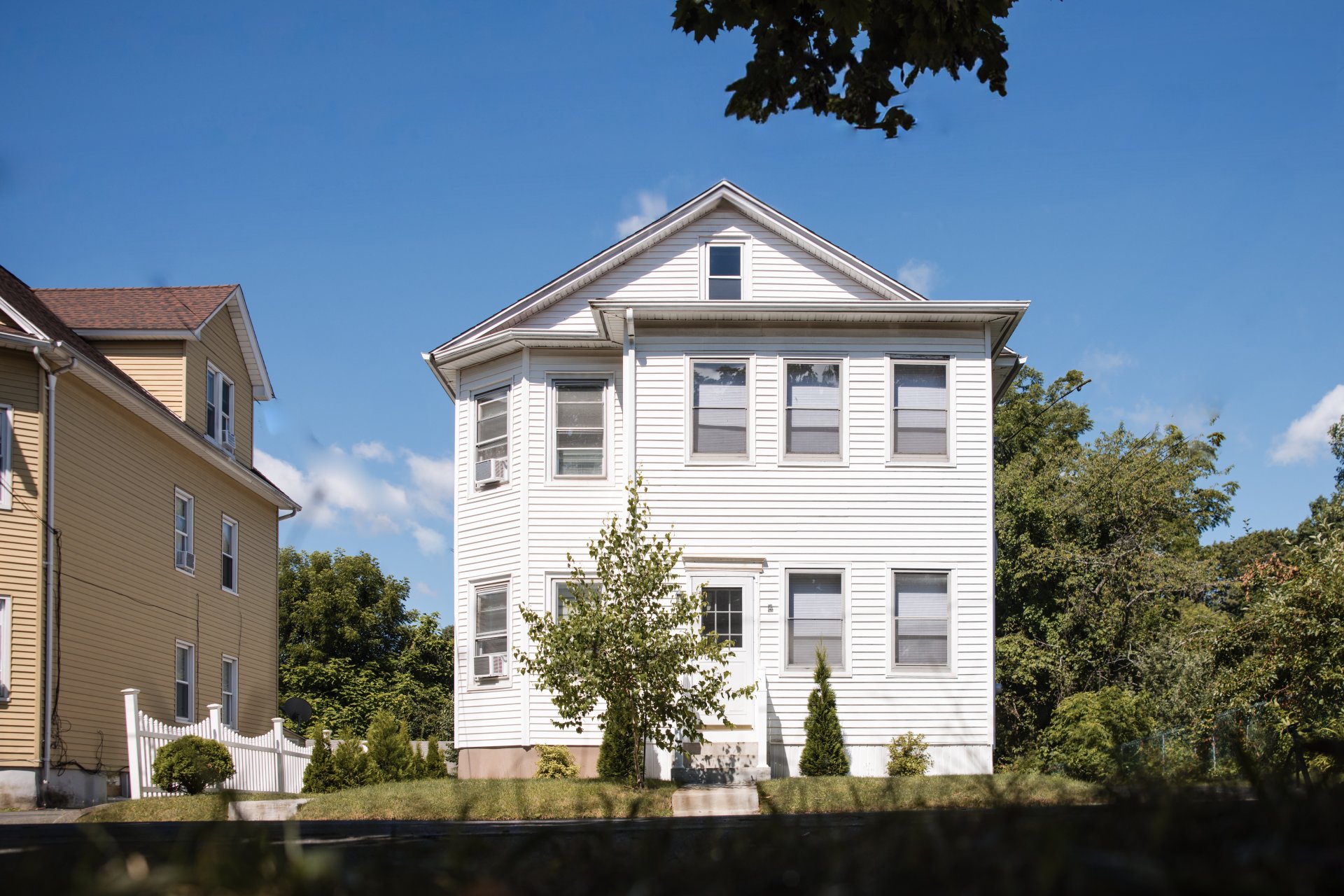  Two-story white house with multiple windows, surrounded by greenery and a neighboring yellow house under a clear blue sky.