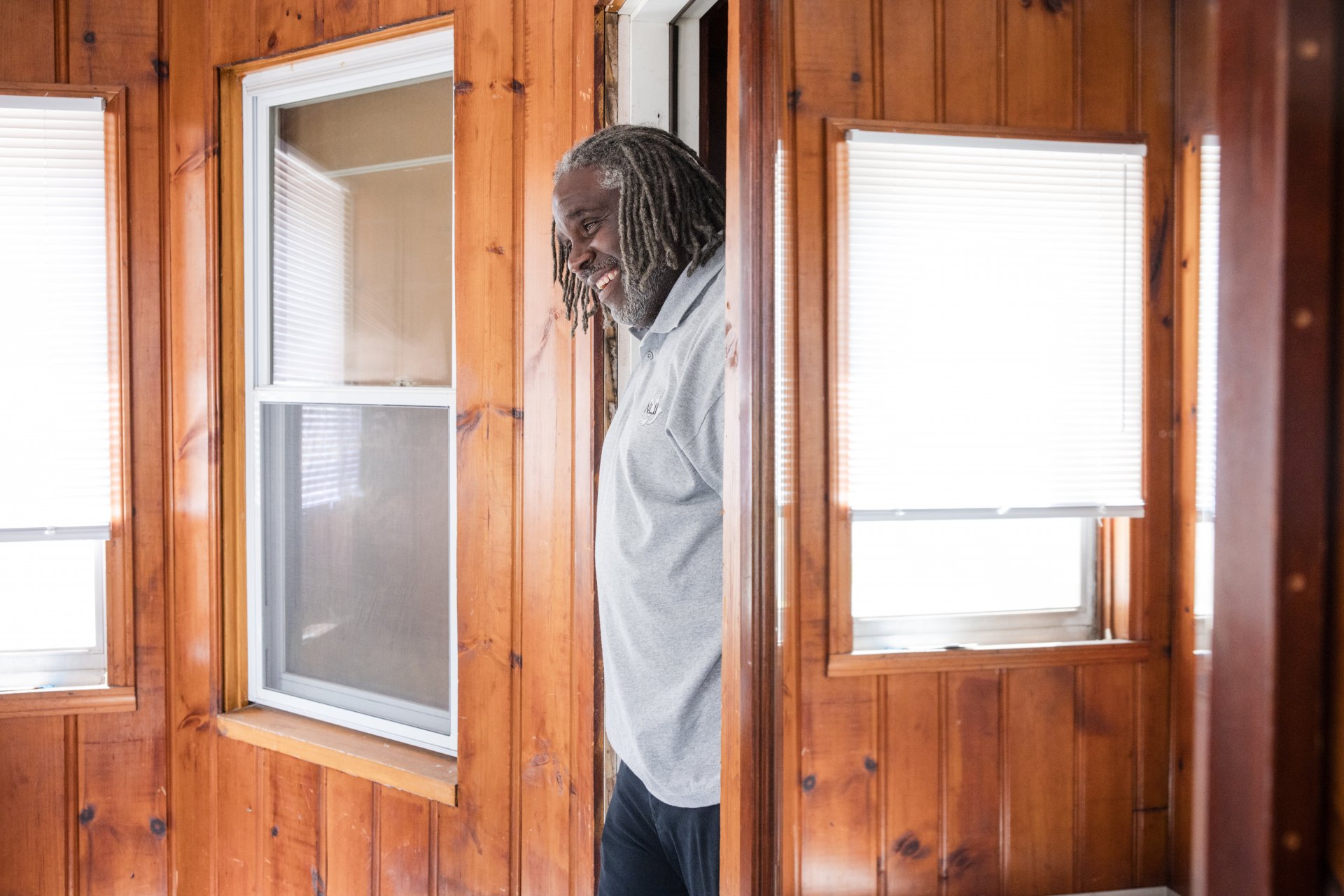 A man with grey dreadlocks and a light shirt stands in a wooden-paneled room, smiling and leaning against a doorway.