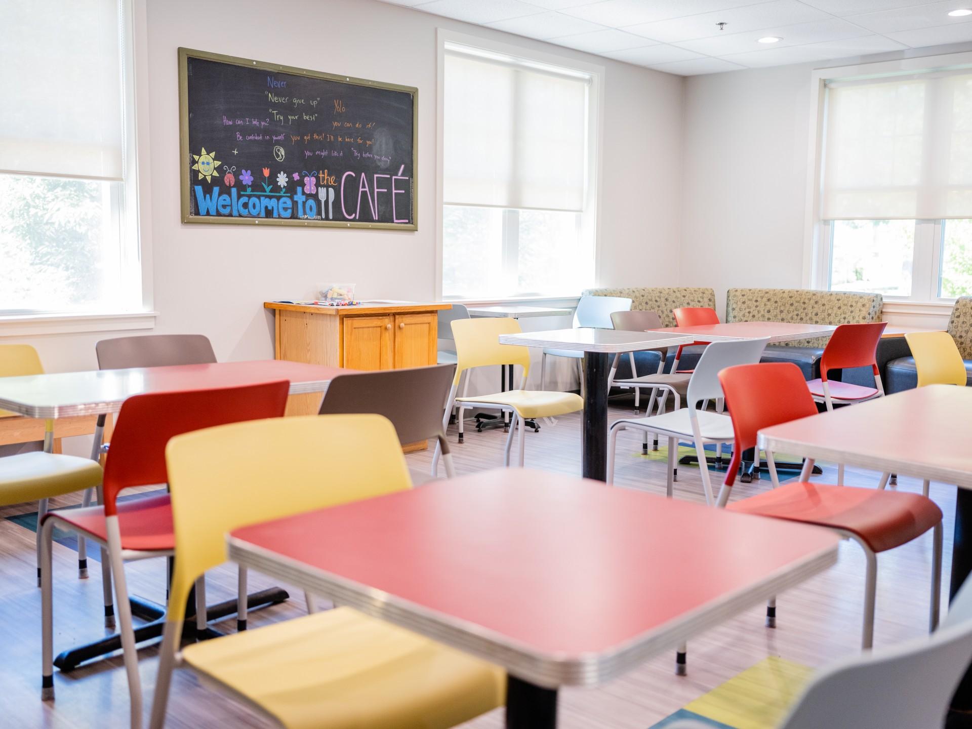 Image of cafeteria room with colorful tables & chairs and a chalkboard on the wall with colorful text reading: Welcome to the Cafe