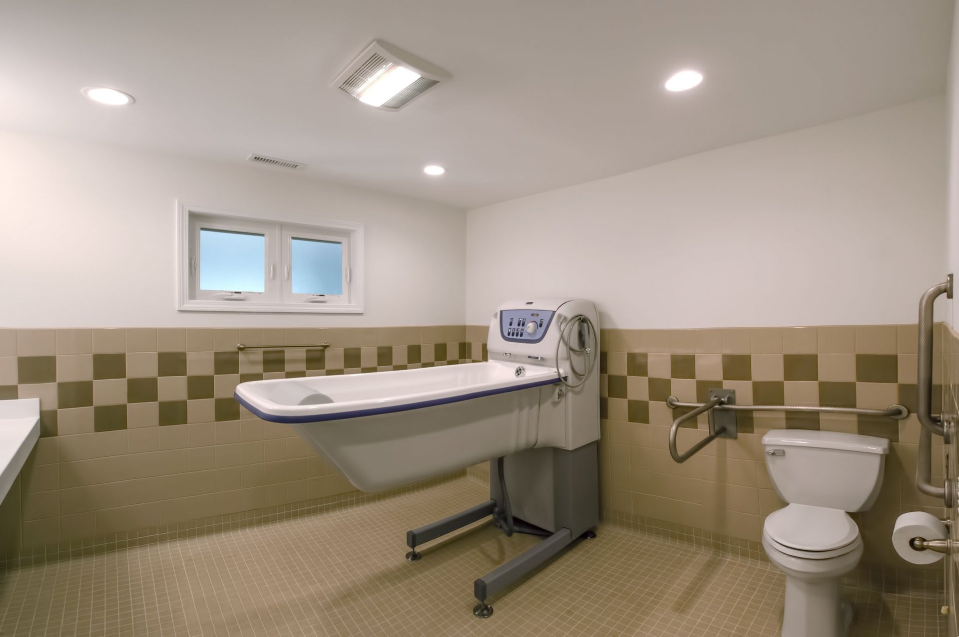  A bathroom with a large medical bathtub, a toilet, grab bars, and beige-tiled walls and floor.